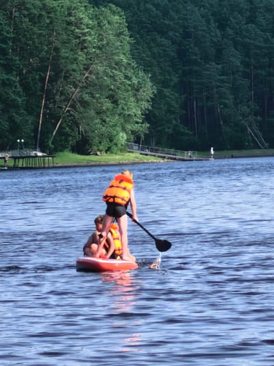 In this image, two individuals are paddleboarding on a calm body of water, possibly a lake, as indicated by the presence of a distant wooden pier and a tree-lined shore. A dense forest forms the backdrop, suggesting a natural, possibly secluded, environment. The water is a deep blue, reflecting the clear sky, and small ripples suggest gentle movement. The scene appears to be set during the daytime, with soft lighting that hints at either early morning or late afternoon.
The two people on the paddleboard are wearing bright orange life jackets, prioritizing safety. One person, standing, is actively paddling with a black paddle, creating a splash of water as they move. The other person is seated on the front of the board. Both individuals appear to be enjoying the activity, with no overt signs of distress. The paddleboard itself is a bright red and white inflatable.
In the background, a wooden pier with multiple supports extends into the water, and several people can be seen on or near it. Further in the distance, more people are visible, some appearing to be walking along the shore or on another platform. The overall atmosphere is serene and suggests a recreational outing in a natural setting. The location is identified as Sysert, Russia. There is no visible text in the image.