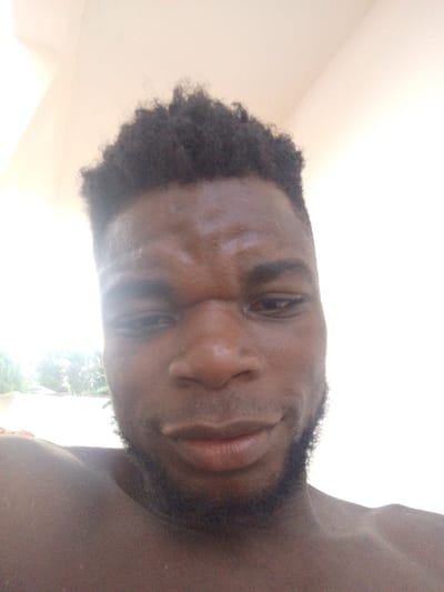 A close-up selfie features a dark-skinned man with a closely cropped, textured hairstyle. He has a short, dark beard. His expression is somewhat serious, with furrowed brows. The background is a bright, indistinct white, possibly a wall or ceiling. The lighting is strong and somewhat unflattering. He appears to be shirtless.