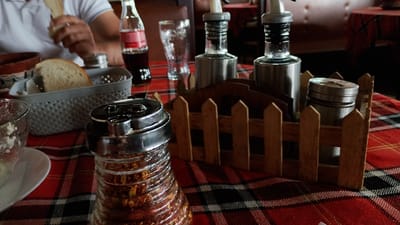 The image depicts a close-up view of a table setting, likely in a restaurant. In the foreground, a clear glass jar filled with red chili flakes is prominently displayed, its ribbed texture visible. To the left, a partially filled glass of what appears to be a white dish or dip sits on a white saucer. Behind these items, a gray, woven basket contains slices of bread. A person's hands are visible to the left, holding a slice of bread as if to eat it. In the background, a Coca-Cola bottle and a clear glass are present, along with two silver cruets or dispensers standing in a wooden holder that resembles a small fence. The table is covered with a red and black checkered tablecloth. The scene is brightly lit, highlighting the textures and contents of the various objects. The overall atmosphere suggests a casual dining environment. The image is likely from Pevtsite, Bulgaria, given the context.