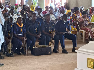 In Kufana, Nigeria, a group of young males in blue uniforms with yellow sashes are seated, holding brass musical instruments, likely preparing for a performance or ceremony. They appear to be a marching band or musical ensemble. Behind them, a diverse crowd of adults, many wearing traditional African attire and headscarves, are also seated, observing the scene. Some are holding papers, possibly programs or religious texts. The environment appears to be an indoor space with earthen floors, suggesting a community hall or a place of worship. The presence of a white, ornate casket in the foreground indicates a solemn occasion, likely a funeral or memorial service. The overall atmosphere is one of respectful gathering and remembrance.