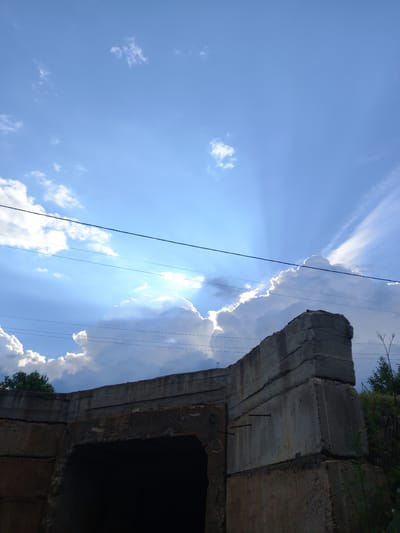 This low-angle, landscape-oriented photograph shows a section of a concrete structure against a bright blue sky. The concrete structure appears to be part of an overpass or bridge, with multiple layered sections visible. The rough texture and visible rebar suggest it is a utilitarian construction. In the sky, scattered white clouds are present, with some large cumulus clouds catching the sunlight. Rays of light are fanning out from behind one of these clouds, indicating a bright, sunny day. Several power lines are strung across the frame, running diagonally and intersecting with the concrete structure and the sky. Vegetation, appearing as green foliage and dry branches, is visible at the top edge of the image, peeking from behind the concrete. The overall scene evokes a sense of an outdoor, possibly rural or semi-urban environment. The time of day is likely midday given the brightness of the sun and the sharp contrast. There is no visible text in the image.