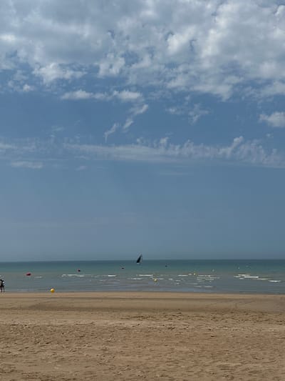 The image depicts a sandy beach with the sea and sky in the background. The sky is partly cloudy with white clouds against a blue backdrop. The sea is calm with gentle waves and appears to be a turquoise color. Several buoys are visible in the water, one yellow and others red. A sailboat with a black sail is also present on the horizon. In the foreground, two people are walking on the beach. The sand is a light brown color and shows some footprints. The scene appears to be a typical day at the beach. This is a view of the Koksijde beach in Belgium.