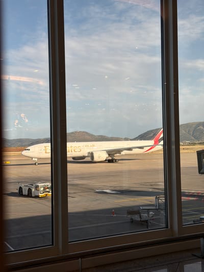 A view from inside a building, likely an airport terminal, looking out through a window onto an airport tarmac. An Emirates aircraft is parked on the tarmac, connected to a jet bridge. The aircraft is white with gold lettering and the Emirates logo. In the background, there are mountains. The sky is blue with some clouds. There is an airport tug vehicle visible near the front of the aircraft. The location is likely Athens International Airport "Eleftherios Venizelos" which is in Spata-Loutsa Municipal Unit, Greece, given the presence of Emirates and the mountainous surroundings.