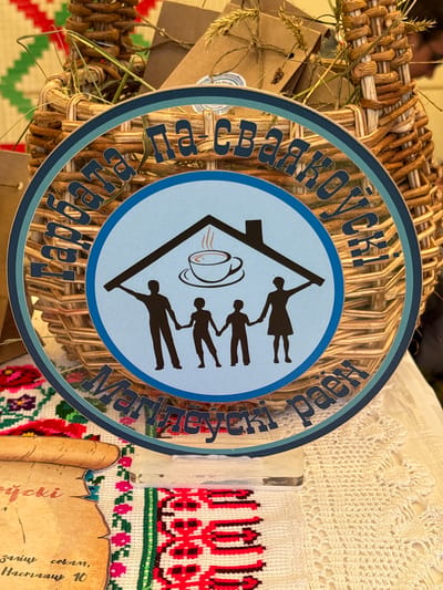 A round blue and teal sign with Cyrillic text is prominently displayed in front of a woven basket. The sign features a stylized house silhouette over a family of four figures holding hands, and a steaming cup of coffee. The basket, filled with dried wheat stalks and small brown packages, is placed on a table covered with a white crocheted doily and a red and white embroidered textile. Below the sign, a scroll-like piece of paper with handwritten text is visible, also resting on the embroidered textile. The scene suggests a marketplace or craft fair setting, possibly in Вяйнянскі сельскі Савет, Belarus, given the presence of traditional Belarusian embroidery and the Cyrillic script.