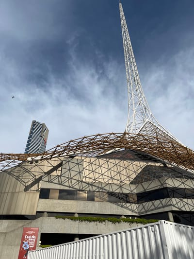 This striking image, set against a bright, partly cloudy blue sky, showcases the unique modern architecture of Federation Square in Southbank, Melbourne, Australia.
The central subject is a dramatic architectural complex. On the left, a tall, slender white lattice-work spire, characteristic of Federation Square's 'shards', tapers upwards. Adjacent to it, a large, curved structure made of brown or gold-colored lattice forms an intricate, organic-looking canopy or facade. This lattice structure casts sharp, geometric shadows onto the building below, which features a complex facade of large, angled glass panels and concrete sections. The geometric patterns of the building's surface are accentuated by these shadows.
In the upper-left, a small silhouette of an airplane is visible against the sky. In the mid-ground, a taller, rectangular building with vertical lines is partially visible behind the main structures. To the far right, a section of a white, corrugated metal structure borders the scene. Greenery can be seen growing along a concrete wall in the lower right.
The bright daylight creates distinct, crisp shadows, indicating a sunny day. A red poster board is partially visible in the lower right, displaying the text "FEELING AFRAID..." and an image of a person in a dynamic pose. The overall scene emphasizes the bold lines and innovative design of the urban landscape.