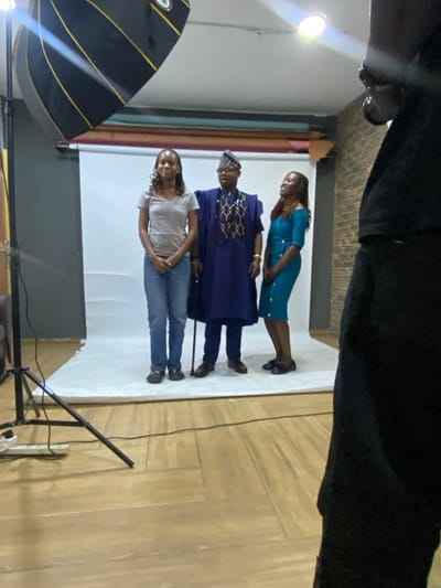 This image captures an indoor photoshoot featuring three individuals against a white seamless backdrop, likely in Animashawun, Nigeria. The photograph is oriented vertically but appears to have been taken horizontally and then rotated 90 degrees clockwise.
In the center stands a man wearing a blue traditional outfit (agbada) with intricate gold and white patterns on the chest, and a matching cap. He appears to be holding a dark cane or staff in his right hand. To his right (viewer's left) is a woman in a light grey t-shirt and blue jeans. To his left (viewer's right) is another woman dressed in a teal or turquoise knee-length dress. All three subjects are standing and facing generally towards the camera, posing for the shot.
The setting includes professional photography equipment. A large black softbox with prominent gold or yellow striped accents is visible on the left side of the frame, indicating a controlled lighting setup. Another bright light source is also visible further to the left. The floor is made of light-colored wood, laid in a herringbone pattern, and a black cable runs across it. Part of a dark object, possibly an arm or shoulder of the person taking the photo or another piece of equipment, is visible at the very top of the frame. The overall impression is that of a professional portrait or group photoshoot in a studio environment. No explicit text is clearly visible on any subjects or equipment.