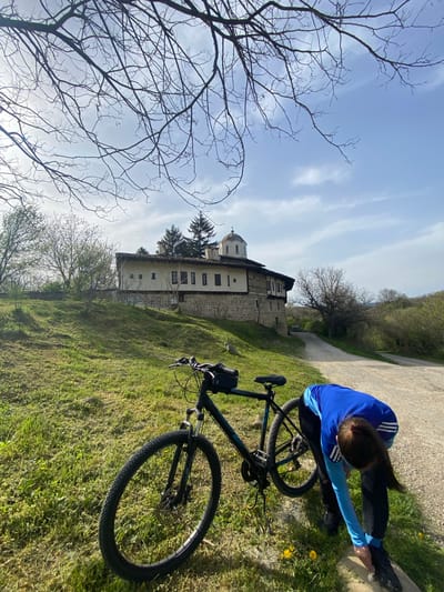 A woman in a blue and white track suit is stretching on a concrete path next to a black mountain bike on a grassy hill. Behind her is a stone building with white trim and a dome, possibly a monastery or church. The location is in Pchelishte, Bulgaria. There are trees with bare branches in the foreground and background. The sky is light blue with wispy white clouds.