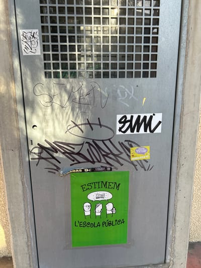 The image depicts a section of a metal utility box on a street in Grao, Spain. The box is covered in various graffiti tags and stickers. A prominent sticker on the lower half of the box is a bright green poster with a cartoon illustration of two figures and text. The poster reads "ESTIMEM L'ESCOLA PÚBLICA" which translates to "We love public school". Above this, there's a sticker with the word "SMI" in bold black lettering on a white background. To the right of the "SMI" sticker, a smaller yellow sticker with Catalan text, "RATIOS RES BAIXES PER ATENDRE MILLOR L'ALUMNAT MOBILITZA'T!", meaning "Fewer low ratios to better serve the students, mobilize!". There is also a torn sticker above these that reads "ENTRADAS 9€". The metal box has a grid of square openings at the top, revealing some inner components. The surrounding environment includes concrete elements and a textured wall, typical of urban street furniture. The lighting suggests it is daytime, with shadows cast across the surface.