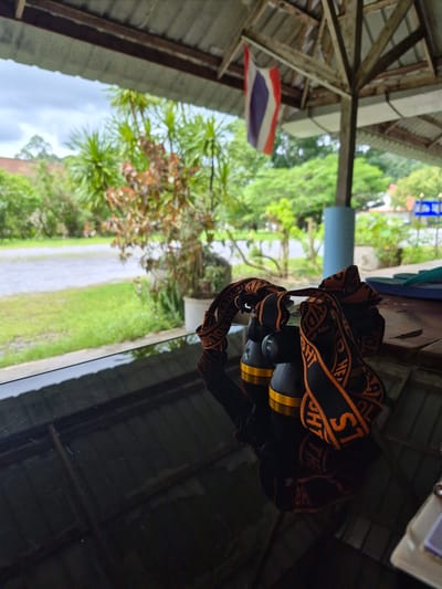 A pair of headlamps with orange and black straps are resting on a glossy black surface, likely a table. The straps appear to be inscribed with text. In the background, through an open-sided structure with a corrugated roof supported by wooden beams, a green landscape with trees and vegetation is visible. A Thai flag hangs from one of the beams. The scene suggests an outdoor setting, possibly a covered patio or a rest stop in a rural or semi-rural area of Phang-nga, Thailand.