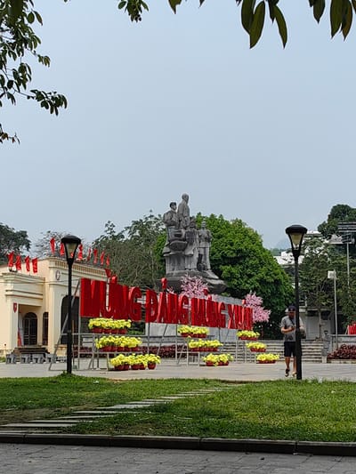 The image shows a public square in Thành phố Hà Giang, Vietnam. In the foreground, there is a patch of green grass with stone pavers leading toward a paved area. In the middle ground stands a large red sign with Vietnamese characters, adorned with pots of yellow flowers. Behind the sign, there is a statue of several figures. To the left, a building with red flags is visible. A man is walking on the right side of the square. Tall trees surround the area. The sky is overcast. The light posts are visible on both sides of the image.