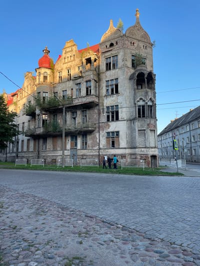 In Sovetsk, Russia, a multi-story, abandoned building with a red-tiled roof stands on a cobblestone street. The building's facade is weathered and decaying, with many broken windows and boarded-up openings. Greenery is visible growing from balconies and the roofline. A white metal fence separates the building from the street. Three individuals, two men and one woman, are walking near the fence. The sky is clear and blue. In the background, other buildings and street signs are visible.