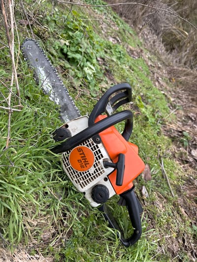 A Stihl 018 chainsaw is depicted lying on a vegetated slope in Radomirtsi, Bulgaria. The chainsaw features an orange engine casing, a white pull-start housing, and black handles. Its metal bar and chain exhibit residue consistent with recent operation. The ground is covered by green grass interspersed with dry vegetation and bare twigs. No individuals are present in the scene.