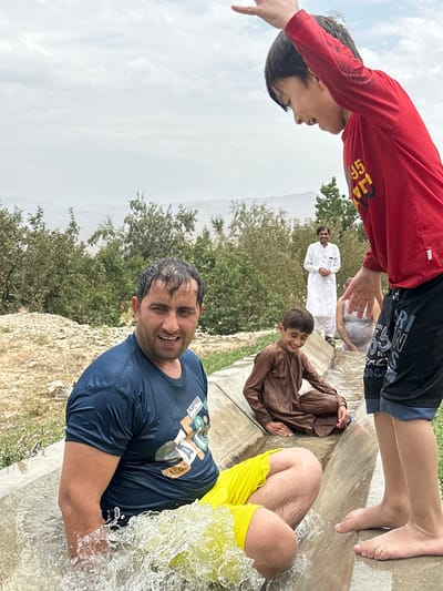 In Kan Mehtarzai, Pakistan, a group of individuals are gathered near a water channel. A man in a dark blue t-shirt and yellow shorts is partially submerged in the water, splashing it with his arm. A young boy in a brown outfit sits further down the channel, smiling. In the background, a man in a white shalwar kameez stands and smiles, while another person is partially visible in the water behind him. The environment appears to be rural, with trees and hills visible in the distance under a cloudy sky.