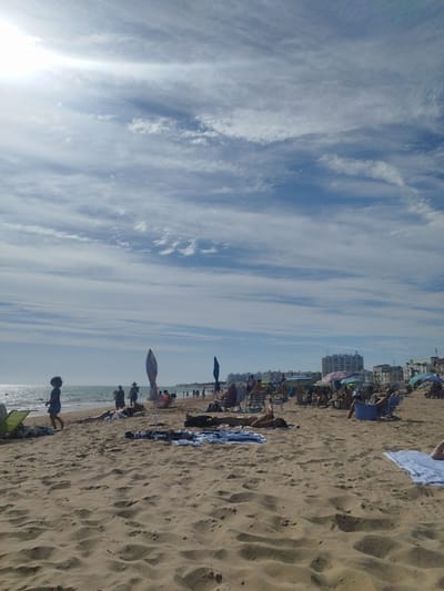 This media file depicts a busy beach scene in Rota, Spain. The image is taken from a low angle on the sand, looking towards the ocean and a populated shoreline. The sun is high in the sky, casting bright light and creating some lens flare in the upper left quadrant of the image. The sky is a vivid blue, with scattered white clouds streaked across it, suggesting a clear and pleasant day.

The foreground is dominated by sandy dunes and textured sand, with a few scattered towels and beach chairs. Several people are visible on the beach, some lounging and sunbathing, while others are walking or standing. In the middle ground, a line of beach umbrellas provides shade for groups of people. The ocean is calm and stretches to the horizon, where the coastline is lined with buildings, suggesting an urban or resort area. Some of the buildings appear to be multi-story apartments or hotels.

The overall atmosphere is one of a relaxed summer day at the beach. People are engaged in typical beach activities such as sunbathing and socializing. The lighting suggests it is daytime, likely late morning or early afternoon. There is no visible text in the image.