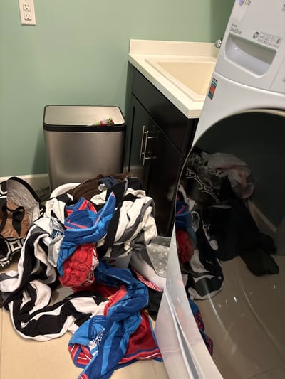A laundry room is depicted with a washing machine in the foreground, a sink and vanity to the right, and a trash can to the left. A large pile of laundry, including towels and blankets, is spread across the floor in front of the washing machine. The room is clean and well-lit, with a pale green wall and tiled floor. This image was taken in Bradenton, Florida, United States.