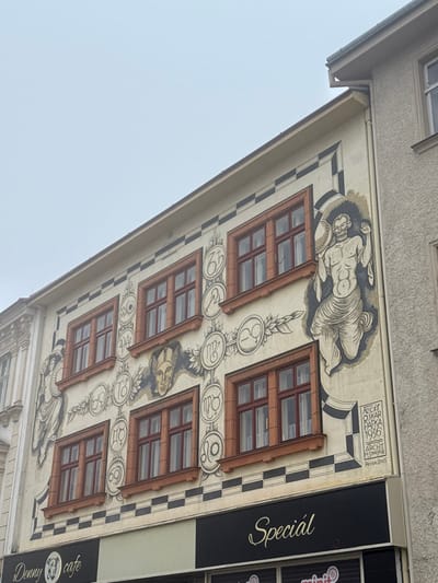The image depicts the sgraffito-decorated facade of a building located at Obroková 3/9 in Znojmo, Czechia, under an overcast sky. The multi-story structure features a light-colored exterior with prominent black and white sgraffito artwork and reddish-brown window frames.

Ten rectangular windows with multi-pane glazing are visible across the two upper stories, five per floor. The facade is extensively decorated with a grid-like arrangement of circular emblems containing astrological or alchemical symbols and Roman numerals (e.g., 'WO', '6S', '69', '18') encircling the windows. A central winged male head is positioned above the lower row of windows.

On the right side of the facade, a large, semi-nude, bearded male figure with drapery around his lower body holds a circular drum or shield in his left hand while raising his right. Below this figure, within a rectangular frame, black text reads: "FECIT OSKAR PAFKA 1936 ARCHT H. SMEJKAL Perov 2005". A partially visible, draped female figure is present on the far left. The sgraffito artwork is framed by black and white checkerboard patterns along the corners and as horizontal bands below the window sills.

The ground floor features a dark commercial fascia. "Denny cafe" with a dog logo is visible on the left, and "Special" is prominently displayed in a cursive font on the right. A portion of "minit" is visible below "Special". An ornate architectural element from an adjacent building is partially seen on the left, while a plain, lighter-colored building with a rain gutter adjoins the right side. The sky above is uniformly grey and overcast.