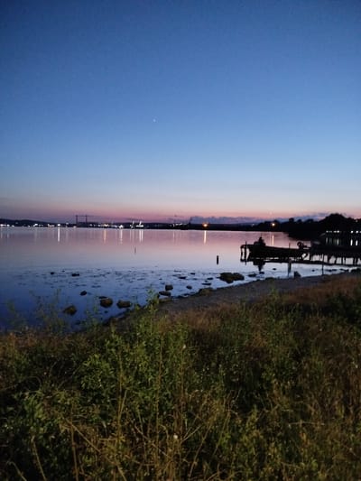 The image depicts a serene twilight scene at the bay of Казашко, Bulgaria. The sky is a gradient of deep blue to soft pink, transitioning towards the horizon. A faint, tiny star is visible in the upper left quadrant of the sky. The calm water reflects the colors of the sky and the distant lights of what appears to be an industrial area or port with prominent smokestacks. On the right side of the image, a wooden pier with a boat docked is silhouetted against the water. The shoreline in the foreground is covered with dry grass, bushes, and scattered rocks. The overall atmosphere is tranquil and peaceful, suggesting the end of a day by the water.