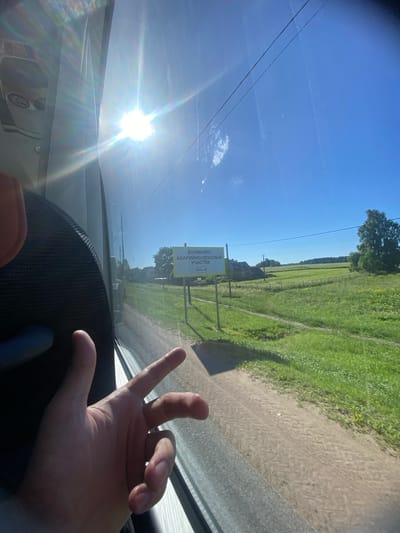 The image shows a view from inside a vehicle, looking out of a window towards a sunny rural landscape. A person's hand is visible in the foreground, gesturing with two fingers extended. Through the window, a bright sunbeam with lens flare illuminates the scene. A road or dirt path runs alongside a lush green field. In the distance, there are several houses, trees, and power lines. A yellow sign with Russian text is prominently displayed on a post, indicating an "EMERGENCY-DANGEROUS SECTION." The sky is clear and blue, with a few wispy clouds. The location is identified as Bol'shoe Teshkovo, Russia.