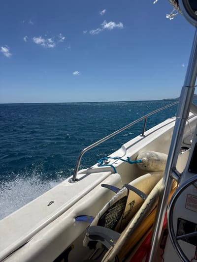 A white powerboat is underway on the open ocean offshore from East Honolulu, United States. The sky above is clear and bright blue with scattered, small white cumulus clouds. The water below is a deep blue, exhibiting surface chop and white spray from the port bow, consistent with vessel movement. A faint, dark landmass is visible on the distant starboard horizon. On the boat's starboard side, a stainless steel handrail runs along the gunwale. A light-colored cylindrical boat fender is secured with a blue rope. Stowed along the starboard interior are multiple recreational boards, including a foil board with a prominent black hydrofoil attachment and at least one light-colored surfboard. A portion of a steering wheel is visible on the far right edge of the frame. No individuals are present in the image.