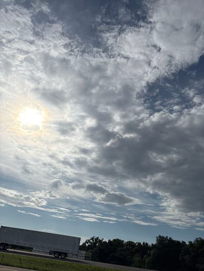 The image shows a view of the sky and a landscape, likely taken from a vehicle on a road in Palmetto, United States. The sky is filled with a mix of dark and light clouds, with the sun peeking through the clouds on the left side of the frame, creating a bright halo effect. A contrail from an airplane is visible in the upper left portion of the sky. In the lower part of the image, a white semi-trailer truck is traveling on a road. Behind the truck, there is a line of dark green trees. A sign is visible on the side of the truck that reads "COMMONS NEW LUXURY APARTMENTS" and a billboard can be seen behind the trees with similar text and an exit number.