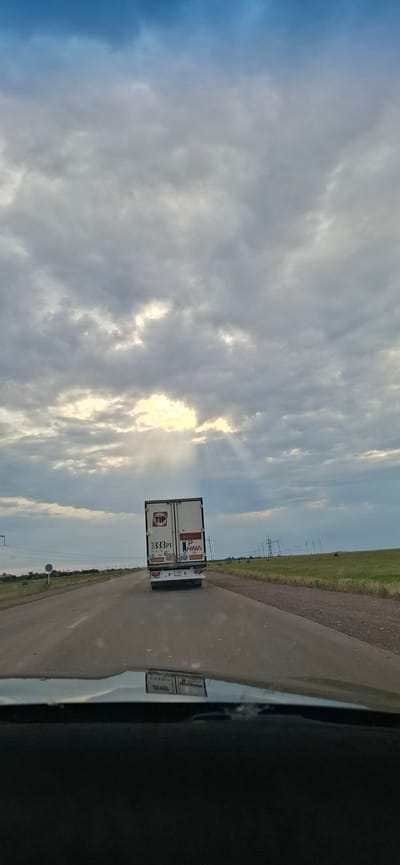 This is a rotated image taken from a vehicle traveling on a road in Жалтыр, Kazakhstan. The primary subjects are a white semi-trailer truck and the sky. The truck is white with several decals on the back. Visible text on the truck includes "TIP", a logo with "HNA", and "333PT". The sky is dramatic, with a mix of dark and light clouds. Sunlight is breaking through the clouds, creating bright rays. The time of day appears to be late afternoon or early evening, given the angle of the sun and the light. The weather seems to be partly cloudy, with the sun intermittently visible. The setting is a rural or semi-rural area, as indicated by the presence of a road, a grassy field to the right of the road, and utility poles in the distance. The overall mood is somewhat serene due to the sky's beauty, but also suggests movement and travel.