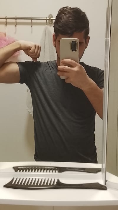 A male subject is depicted in a mirror selfie taken within an indoor environment, likely a bathroom. The specific city of Neunkirchen, Germany, is not identifiable from the visual information. The subject, seen from the chest up, has dark hair and is wearing a dark gray short-sleeved t-shirt. His face is largely obscured by a light-colored smartphone held in his right hand. His left arm is flexed, with the hand forming a fist, showcasing his bicep.

The background consists of a light-colored wall. A horizontal metal towel bar with four hooks is mounted on this wall. A pink bath pouf hangs from the leftmost hook, and a folded white towel hangs from the second hook from the left. Below the mirror, a white shelf or countertop is visible. Two dark-colored combs are positioned horizontally on this surface: one with widely spaced teeth closer to the viewer, and another with finer, more numerous teeth positioned behind it. Artificial overhead lighting illuminates the scene.