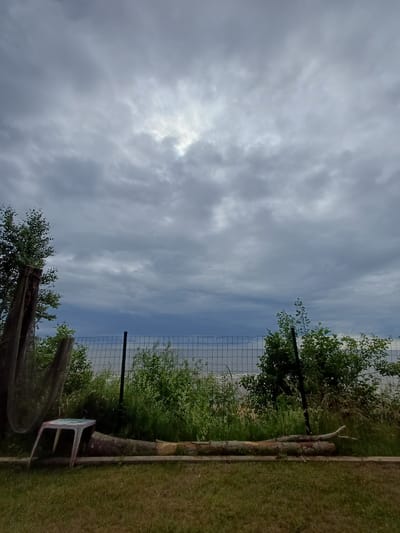 A cloudy, overcast day is depicted in this outdoor scene. The sky is filled with heavy gray clouds with some brighter patches where the sun is attempting to break through. In the foreground, a patch of mown grass leads to a wooden log placed horizontally. To the left of the log, an overturned, light blue plastic stool rests on the ground. Further back, a wire mesh fence runs horizontally across the image, separating the foreground from a strip of lush green vegetation and trees. Beyond the vegetation and fence, a body of water, likely a lake or sea, is visible under the dusky sky. The location is identified as Vārzas, Latvia.