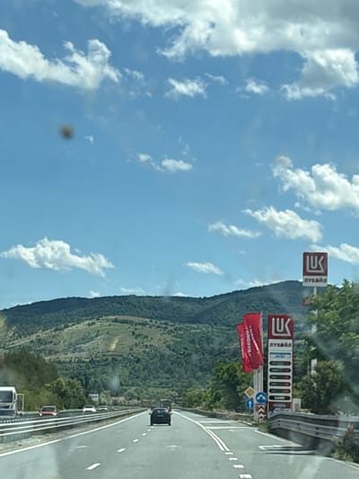 The image captures a view from a car driving on a highway, likely in or near Studena, Bulgaria, given the presence of a Lukoil gas station. The sky is blue with scattered white clouds. In the foreground, the highway is clearly visible with multiple lanes and other vehicles. To the right, a tall Lukoil sign displays fuel prices and logos. Red banners are also visible near the gas station. In the background, a lush green mountain range dominates the landscape, with trees and some terraced areas. The overall scene suggests a journey through a scenic, rural or semi-rural area.