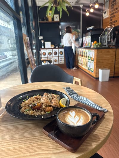 A medium shot captures a table in a cafe in Tha Chana, Thailand, with a meal and coffee. In the foreground, a black plate holds a dish of rice topped with grilled salmon and garnished with chopped green onions and sesame seeds, accompanied by a lemon wedge. Next to the plate is a dark mug of coffee with latte art, resting on a wooden coaster. A grey and white patterned napkin is folded beside the plate. The table itself is light wood with visible grain. In the background, a woman in a white shirt and dark pants stands behind a counter, which is adorned with various food posters and illuminated by ceiling lights. A shelf above the counter displays items including a potted plant and framed pictures. A trash bin is visible near the counter. Through a large glass window to the left, reflections and signs indicate the cafe's location. The overall environment suggests a casual dining setting.