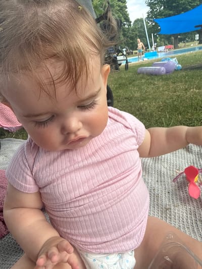 The media file is an image of a baby lying on a blanket outdoors. The baby is wearing a pink ribbed onesie and a diaper. The baby's eyes are closed, and they appear to be sleeping or resting. In the background, a swimming pool and trees are visible, suggesting the setting is a park or backyard with a pool. There is also a blue umbrella and some inflatable toys. The lighting suggests it is daytime. The overall mood of the image is peaceful and relaxed, with the baby appearing content. There is no visible text in the image.
