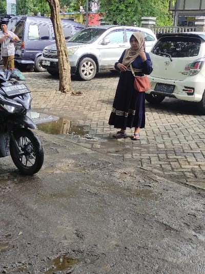 The image depicts a daytime outdoor scene in Kecamatan Asembagus, Indonesia, showing a street or parking area. The ground features two distinct sections: a lighter paved sidewalk made of rectangular blocks on the left, and a darker, rough, wet, and muddy concrete or asphalt surface with puddles on the right, indicating damp conditions possibly from recent rain.
A woman is prominently featured standing on the paved area, facing slightly to her right. She wears a long dark blue dress with patterned trim, a matching long-sleeved top, and a light beige headscarf (hijab). A light orange shoulder bag is slung across her body. Her expression is neutral, possibly with a slight smile. Behind her, a white hatchback car is parked, with its rear visible. The license plate "P 7288 SG" is partially legible, and a "Toyota" emblem is visible on the trunk.
Further along the paved area, a silver SUV or crossover is parked, showing its side profile and a wheel with a "Honda" logo. Behind this, a dark-colored van or MPV is also visible. A tree trunk stands in the mid-ground, separating the paved section from the street.
On the rough, wet ground to the right, a black motorcycle is parked, with its front wheel and part of its body visible. Its license plate, "P 6768 FS", is partially readable. In the lower left background, near the motorcycle and a tree, a man in a light shirt and trousers, possibly wearing a cap, is standing. Green foliage is visible in the far left background. The scene captures an everyday moment in an urban or semi-urban setting.