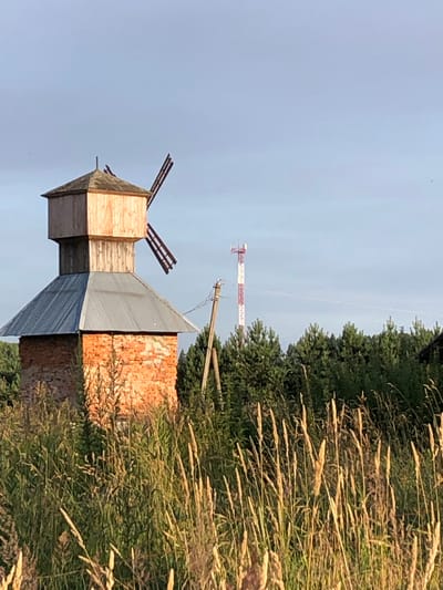 In Makeikha, Russia, a wooden windmill with a brick base stands in a field of tall grass. The windmill has a conical metal roof and wooden blades. In the background, a tall cell tower with red and white bands is visible above a dense line of green trees. The sky is a pale blue.