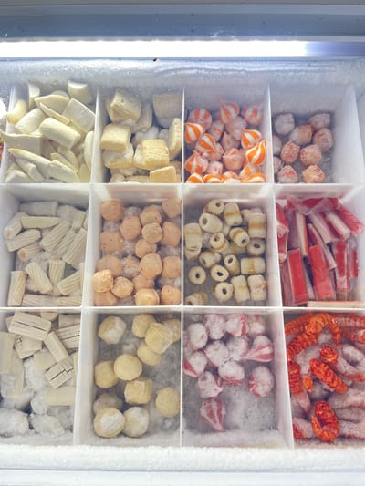 The image shows a close-up, overhead view of a freezer display case filled with various frozen food items, divided into a grid of compartments. The compartments contain items such as white, cylindrical pieces of food in the top left, cubed white pieces in the top middle, striped orange and white rounded items in the top right second from left, and reddish-orange spherical items in the top right. The second row from the top features white, cylindrical pieces with indentations in the left compartment, light brown spherical items in the middle left, white rings with brown stripes in the middle right, and red and white stick-like items in the right compartment. The bottom row contains white, rectangular items with grid patterns in the left compartment, light yellow spherical items in the middle left, red and white striped spherical items in the middle right, and red, shrimp-like items in the right compartment. The freezer is covered in a layer of frost. The location is the city of Pasar Paloh, Indonesia.