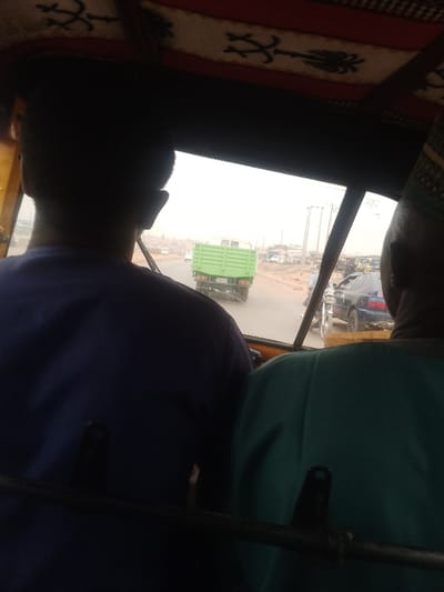 This image, situated in the city of Lapai, Nigeria, depicts an interior view from a vehicle, likely a tricycle, looking forward onto a road with active traffic. In the immediate foreground, the backs of two individuals are visible. The person on the left wears a dark blue garment, and the person on the right wears a teal garment and a patterned cap. A black horizontal bar, part of the vehicle's interior structure, crosses the lower frame. The vehicle's ceiling is lined with a white fabric featuring dark, stylized patterns against a red border.
Through the windshield, a light green box truck or dump truck is observed directly ahead, receding into the distance on a dusty or unpaved road. To the right of the truck, a dark blue sedan with prominent taillights and dual exhaust tips is visible, alongside other partially obscured vehicles, including a motorcycle. The background features various low-rise structures and buildings lining the roadside, accompanied by utility poles carrying multiple wires. The sky appears overcast or hazy, contributing to diffuse lighting.