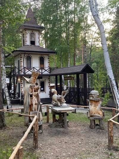 In the city of Tal'tsy, Russia, a picturesque outdoor scene unfolds, featuring a charming, whimsical architectural structure reminiscent of a fairytale tower, nestled amongst lush green trees. The tower, a two-story building with a conical roof, is adorned with white walls and brown brick accents, complemented by arched windows and a wooden balcony.

In the foreground, several wooden sculptures add to the enchanted atmosphere. To the left, a tall, imposing figure carved from wood stands proudly, clad in a flowing cape and a crown, holding a sword. In the center, a smaller sculpture depicts a creature or animal resting on a table, surrounded by intricate carvings. To the right, another wooden sculpture resembles an owl or a gnome, perched on a wooden base, with visible text carved into its surface.

A rustic wooden fence, made of rough-hewn logs, encloses parts of the scene, guiding visitors along a gravel path. The ground is covered with a mix of gravel and fallen leaves, with patches of grass peeking through. The surrounding environment is dominated by tall trees, predominantly birch trees with their distinctive white bark, creating a serene and natural backdrop. The lighting suggests it is either early morning or late afternoon, with soft sunlight filtering through the leaves, casting gentle shadows. There are no people visible in the frame, leaving the viewer to imagine the stories and magic that might unfold in this enchanted setting.