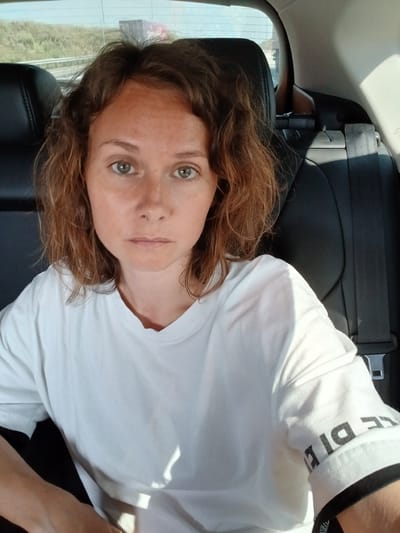 A woman with light brown, wavy hair and green eyes is pictured from the chest up, seated in the back of a car. She is wearing a white t-shirt with black lettering on the sleeve. Her expression is neutral. The background shows the interior of the car, including a black leather seat and a seatbelt, and through the rear window, a blurred view of trees and a road. The overall lighting suggests it is daytime.
