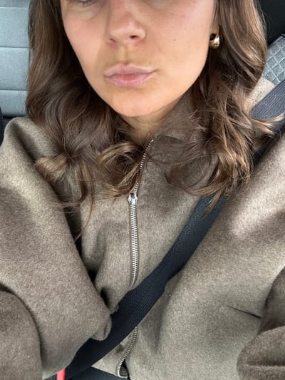 A close-up image depicts a person, identified as female, seated in a vehicle located in Mougins, France. The subject's face is partially visible, showing the nose, mouth with slightly pouted lips, and chin, framed by medium brown, wavy hair. A gold-toned hoop or stud earring is visible on the right earlobe. The individual is wearing an olive-brown, textured fabric jacket, secured with a silver-colored zipper that is partially closed. A black seatbelt crosses diagonally over the subject's chest and right shoulder. The background reveals a dark gray, quilted upholstery consistent with a vehicle's interior, likely a headrest or seat.