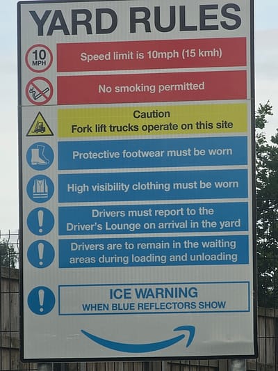 The image depicts a rectangular sign titled "YARD RULES". The sign is divided into vertical colored sections. The first section is red and states "Speed limit is 10mph (15 kmh)" with a circular speed limit sign. The next red section states "No smoking permitted" with a smoking prohibition symbol. Following this is a yellow section with "Caution Fork lift trucks operate on this site" and a warning sign with a forklift. The remaining sections are blue, stating: "Protective footwear must be worn" with a boot symbol; "High visibility clothing must be worn" with a vest symbol; "Drivers must report to the Driver's Lounge on arrival in the yard" with an exclamation point in a circle; "Drivers are to remain in the waiting areas during loading and unloading" with an exclamation point in a circle; and finally "ICE WARNING WHEN BLUE REFLECTORS SHOW" and a blue arrow. The sign is posted in Garden City, United Kingdom. The sign lists essential rules for a specific yard or site.