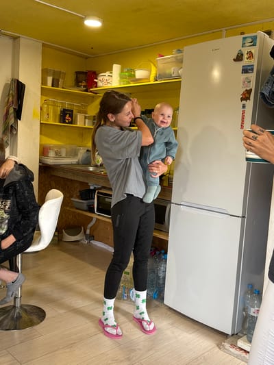 In Dulepovo, Russia, a woman in a grey t-shirt and black leggings is holding a smiling baby in a blue onesie. The woman is standing in a kitchen with yellow walls and shelves filled with various items including food containers, pots, and bottles. A white refrigerator stands to the right of the woman, adorned with several magnets. To the left of the woman, a person wearing a black hoodie is seated on a white stool. The floor is made of light-colored wood. The overall scene depicts a casual indoor gathering.