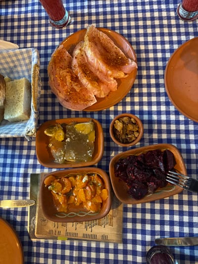 A variety of food items are arranged on a blue and white checkered tablecloth. Plates and bowls contain thinly sliced meat sprinkled with spices, dark red beets, minced meat, aspic, and carrots. A basket of bread sits near a bottle of red sauce. Utensils like forks and knives are present. A wooden menu board is also visible on the table.