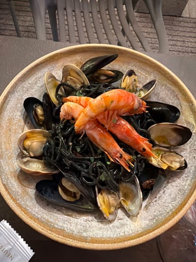 A close-up, overhead shot captures a rustic, ceramic bowl filled with squid ink pasta and an assortment of fresh seafood. The dark, glistening pasta serves as a dramatic backdrop for plump, pink shrimp and various shellfish, including mussels and clams. The clams are partially open, revealing their succulent interiors, and the mussels, with their dark, iridescent shells, are nestled amongst the pasta. The shrimp are prominently featured, their vibrant orange contrasting with the dark pasta. A scattering of finely chopped green herbs adds a touch of freshness to the dish.

The bowl itself has a textured, speckled surface with a warm, earthy tone and a slightly raised, golden-brown rim, suggesting a handmade or artisanal quality. The lighting appears to be warm and somewhat dim, possibly from an indoor setting or evening light, casting soft shadows and highlighting the glossy textures of the food.

In the background, to the left, a textured, grey wall or partition with a grid-like pattern is visible, possibly a decorative element of the restaurant's interior. The setting suggests a relaxed dining experience. To the lower right, a small, white packet with the word "NEUTRA" partially visible is placed on a dark surface, possibly a condiment or napkin wrapper. The overall impression is one of a delicious, well-prepared seafood dish presented in an appealing and rustic manner.