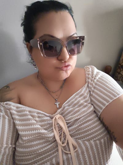 A woman with dark hair pulled back is wearing large, light-colored sunglasses and a beige and white striped off-the-shoulder top. She has on a necklace with a cross pendant and small earrings. She is making a kissing face. The background appears to be a wall. There is no indication of the city of Komotini, Greece.