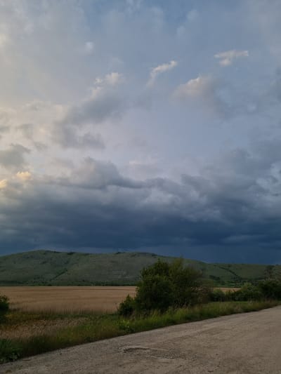 The image shows a view in Aldomirovtsi, Bulgaria. On the right side is a gravel road with green vegetation growing along its edge. Adjacent to the vegetation is a field of harvested crops. In the background is a green hillside with the text "1995-2020 TUK SME ZAEDNO" written in white. The sky is cloudy, with a mix of light and dark gray clouds.