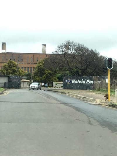 A roadside view features a large "Kelvin Power" sign in dark stone, a traffic light, and a street. A large, industrial brick building with smokestacks stands in the background, partially obscured by trees. A white car is visible on the street near the sign. The sky is overcast. The road appears wet, possibly from rain.