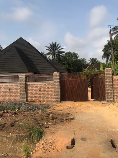 This is a daytime photograph taken in Ihiala, Nigeria, depicting the exterior of a residential property and its immediate surroundings. The primary subject is a house featuring a dark gray, multi-faceted pyramidal roof constructed from shingle-like material, partially visible behind a perimeter fence.
The fence is composed of reddish-brown and gray brickwork columns and base sections, topped with white or cream plaster and a horizontal brown metal railing. An entrance gate, made of vertical corrugated brown metal, occupies the center-right of the fence line. The right leaf of this double gate is partially open, revealing a dirt pathway leading into the property.
The foreground consists of a reddish-brown dirt road or path. On the left side of this path, a concrete drainage culvert or gutter opening is present, partially filled with earth and small rocks. Sparse green grass and weeds grow in patches along the culvert and on the adjacent ground. A small pile of dark gray gravel and some scattered refuse are visible on the ground to the left of the gate, inside the fence line.
In the background and surrounding the property, numerous tall palm trees and other dense green tropical foliage are evident under a light blue sky with scattered white cumulus clouds. A weathered utility pole, potentially wooden, with a horizontal cross arm is visible on the far right edge of the frame, partially obscured by trees. No individuals are present, and no active interactions or ongoing actions are depicted in the scene.