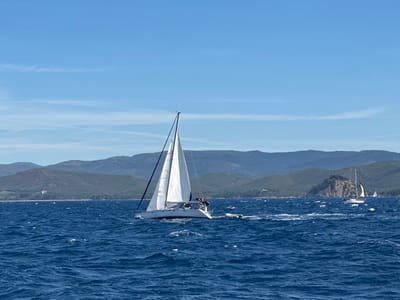 This is a photograph of a bright, sunny day at sea, with a clear blue sky and scattered thin clouds. In the foreground, the deep blue sea is choppy with whitecaps. A white sailboat with a partially furled sail is sailing on the right side of the frame. The boat appears to be moving forward, leaving a wake behind it. A few people are visible on the deck of the sailboat. Further in the distance, a coastline with green, rolling hills and mountains is visible. The coastline is dotted with some buildings. There is another sailboat in the far distance on the right. The image depicts a nautical scene possibly near the coast of Bormes-les-Mimosas, France, known for its beautiful Mediterranean coastline.