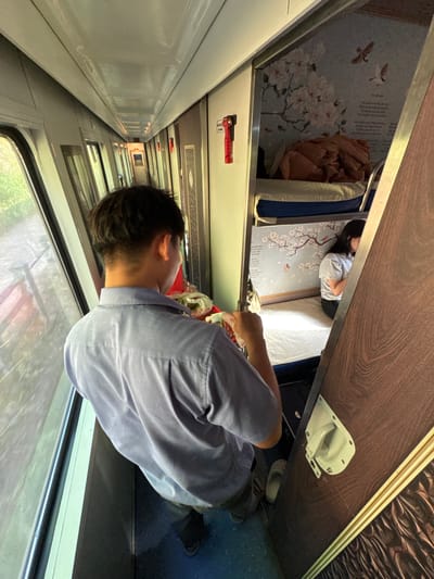 The image depicts the interior of a train compartment in Châu Ổ, Vietnam. A man wearing a light blue collared shirt is standing, facing away from the camera and looking towards a bunk bed area. He appears to be holding a plastic bag with some food items. The bunk beds are visible, with one occupied by a woman in a striped t-shirt. The compartment walls are decorated with floral wallpaper and images of birds. The train carriage has large windows on the left, offering a view of greenery outside. The overall scene suggests travel or accommodation within a train.