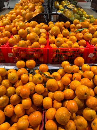 This image captures a vibrant, close-up view of a large display of fresh oranges, likely in a supermarket or fruit market in Kelapa Dua, Indonesia. The scene is dominated by piles of round, orange-colored citrus fruits, many of which still have small green stems or leaves attached, suggesting their freshness.

The oranges are primarily displayed in red plastic crates, stacked or arranged to showcase the abundance of the produce. In the background to the left, another section of the display holds green fruits, possibly limes, lemons, or green mandarins, indicating a variety of fresh produce available. The lighting appears to be artificial overhead light, common in indoor retail settings.

A white price tag or label with the number "70" is visible on one of the red crates, potentially indicating a price per unit (e.g., per kilogram or per item) or a product code. The perspective is a slightly downward-looking shot, emphasizing the sheer quantity and natural texture of the fruits. The overall impression is one of a well-stocked and inviting fresh produce section, highlighting the rich color and quality of the citrus fruits. No people are visible in the frame.