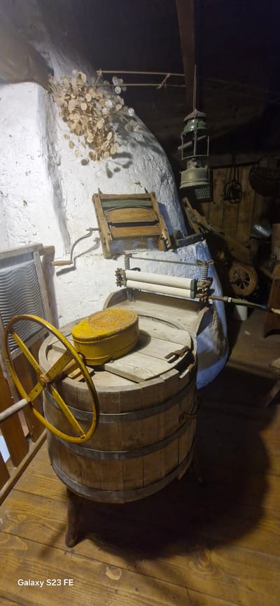 The image captures a scene within the Pakruojis Manor complex in Lithuania, specifically an interior space resembling a historical utility or laundry area. The lighting is dim, casting long shadows, and the primary subjects are antique laundry appliances and a wooden barrel.

In the foreground, a large wooden barrel serves as a central object. It has metal hoops reinforcing its structure and a wooden lid. A prominent yellow, round metal object with a raised lid rests on top of the barrel's lid. To the left of the barrel, a large, yellow, spoked wheel is attached to a handle, suggesting a manual operation mechanism.

Behind the barrel, mounted on a white, textured wall, is an old wooden washboard with a ribbed metal surface on one side. To its right, another wooden washboard is visible. Above the washboard, a vintage oil lamp with a green top hangs from a wooden beam.

Further in the background, a wooden machine with two rollers is positioned. This appears to be a wringer, likely used in conjunction with the washboards for squeezing water out of laundry. The entire setup evokes a sense of a past era, showcasing historical methods of laundry care. The environment appears to be rustic, with wooden floors and rough-textured walls, consistent with the historical context of a manor estate. The presence of dried plant material hanging on the wall adds to the aged and somewhat preserved atmosphere.