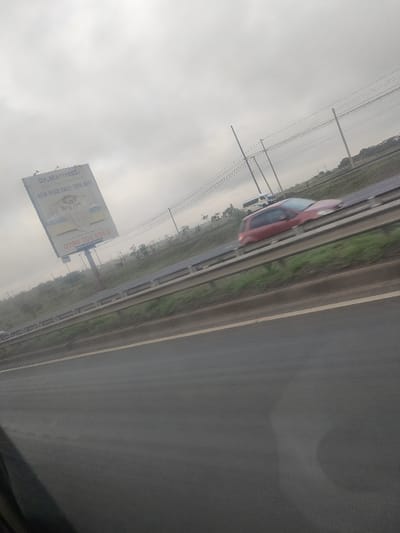 The image depicts a roadside scene on a highway in Gatongora ward, Kenya, under an overcast sky. In the foreground, the edge of a dark vehicle interior is visible in the lower left. The paved asphalt highway features a white solid line on its right edge, bordering a grassy embankment. A metallic multi-bar guardrail runs parallel to the highway along this embankment.
On the raised grassy embankment, a large billboard is prominently displayed, advertising "Dr. Mattress" with "NEW YEAR SALE: 20% OFF!" and a phone number (0790 202 991/2). The billboard design includes an image of a bed with white bedding. Multiple wooden utility poles with overhead power lines stretch into the distance along the roadside, following the curve of the highway.
Vehicles are in motion on the highway, indicated by motion blur. A red hatchback car is visible in the lane adjacent to the guardrail, moving from right to left across the frame. Further in the background, a white van or SUV is also in motion on the highway. Distant vehicles are faintly visible on the highway in the far background. The overall environment suggests a rural or semi-urban highway setting.